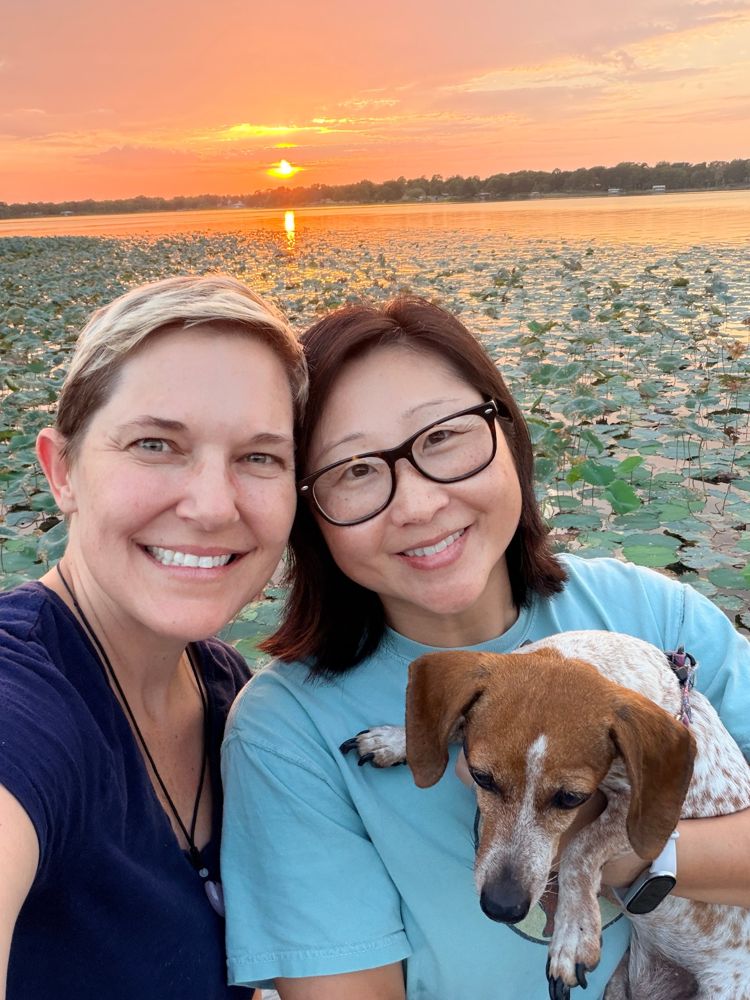 Annie M. Henderson and her wife Sam at sunset by a lake, holding their dog Skeeter